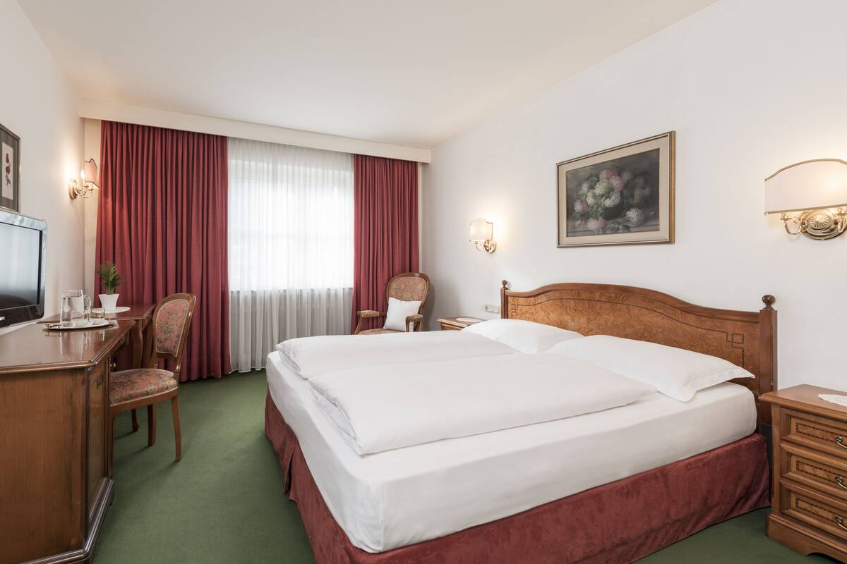 Habitación de hotel con cama, escritorio con televisor, silla, mesita de noche y lámpara de pared.