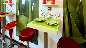 Interior de un restaurante con taburetes de bar verde y rojo, una mesa verde con platos y una pared decorada con motivos florales.