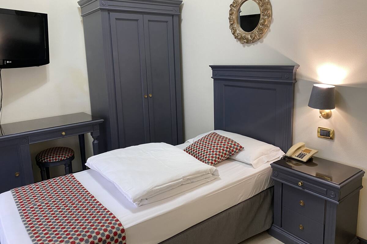 A neatly made bed with white linens and a red and white patterned blanket, a bedside table with a lamp, a telephone, and a mirror on the wall.