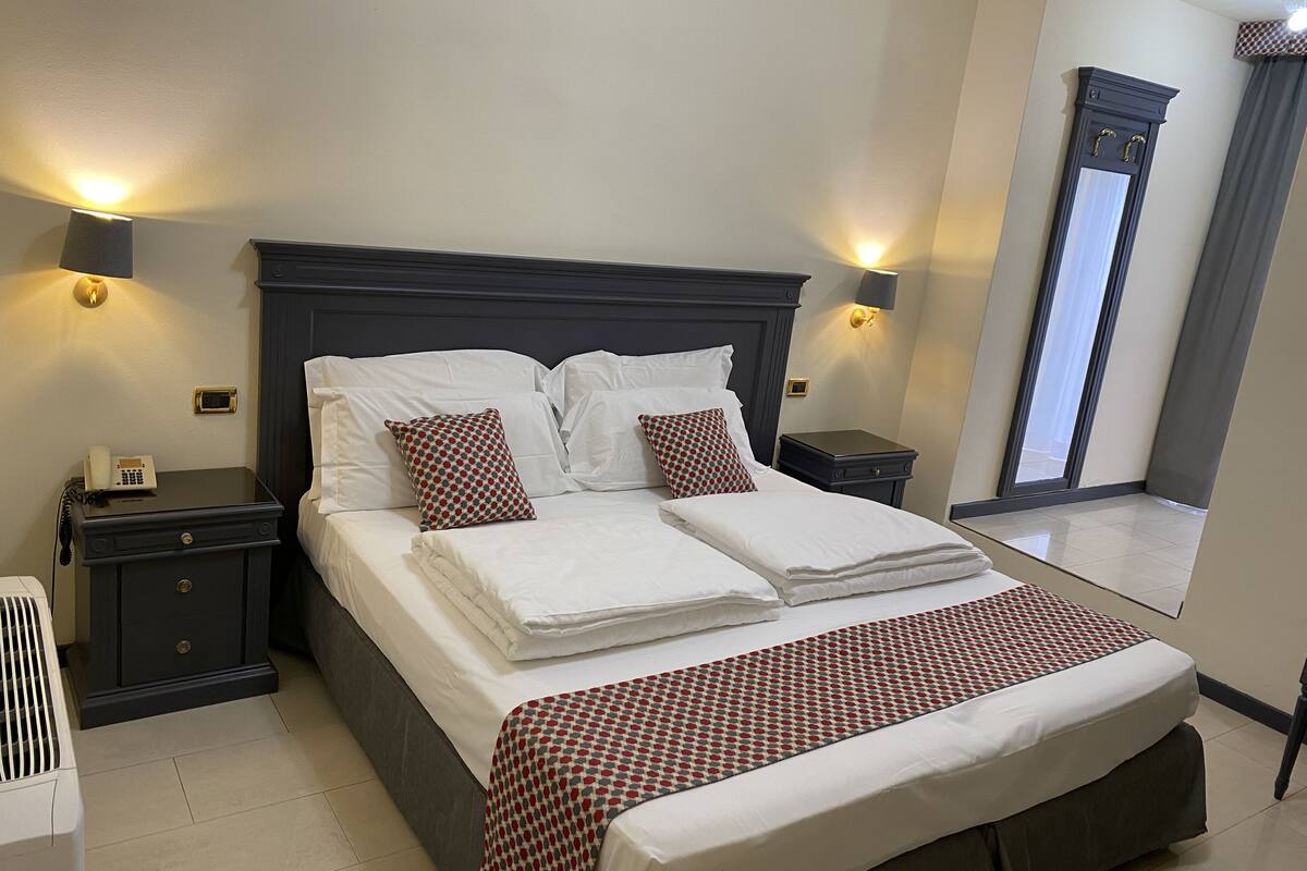 A neatly made bed with white linens and red and white patterned pillows, two bedside tables with lamps, a telephone, and a mirror on the wall.