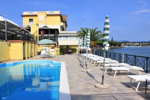 Zona de piscina con sillones de descanso y sombrillas, un edificio con letrero que indica un hotel, y vista al mar.