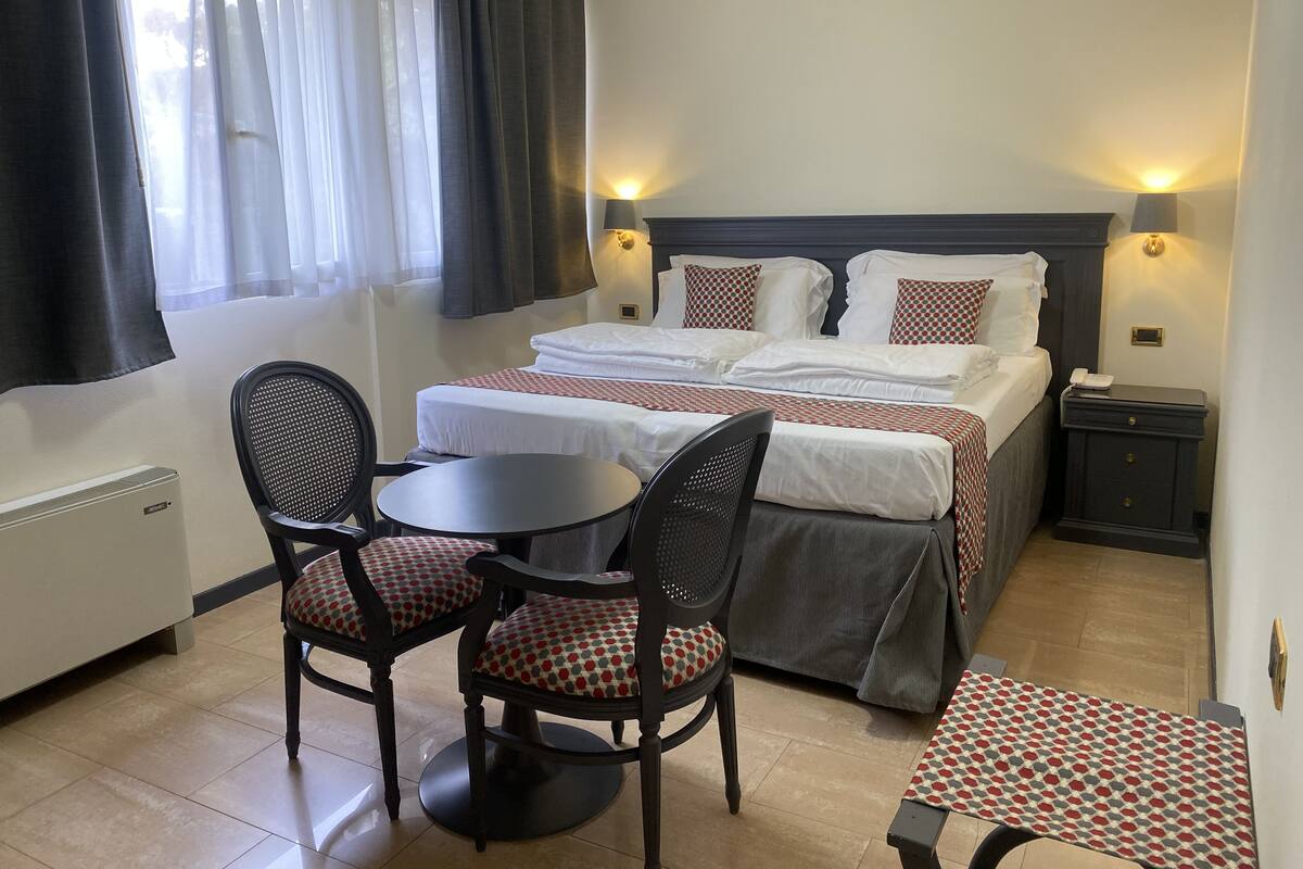A hotel room with a bed, two bedside tables, a chair, a small round table, and a desk with a chair.