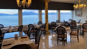 Um restaurante com mesas e cadeiras, grandes janelas com vista para o mar e montanhas, e lustres.