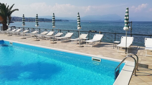 Área à beira da piscina com espreguiçadeiras e guarda-sóis, com vista para um corpo d'água e montanhas ao longe.