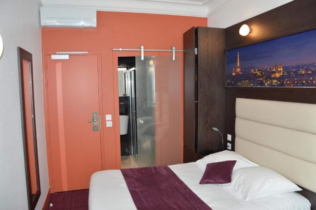 A hotel room with a bed, a view of the cityscape, and a door leading to a bathroom.