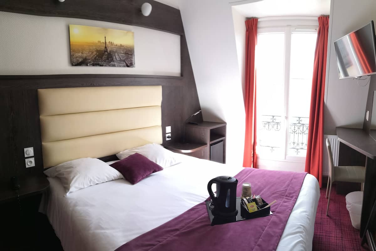 A hotel room with a large bed, a nightstand, a desk, a TV, and a balcony with red curtains.