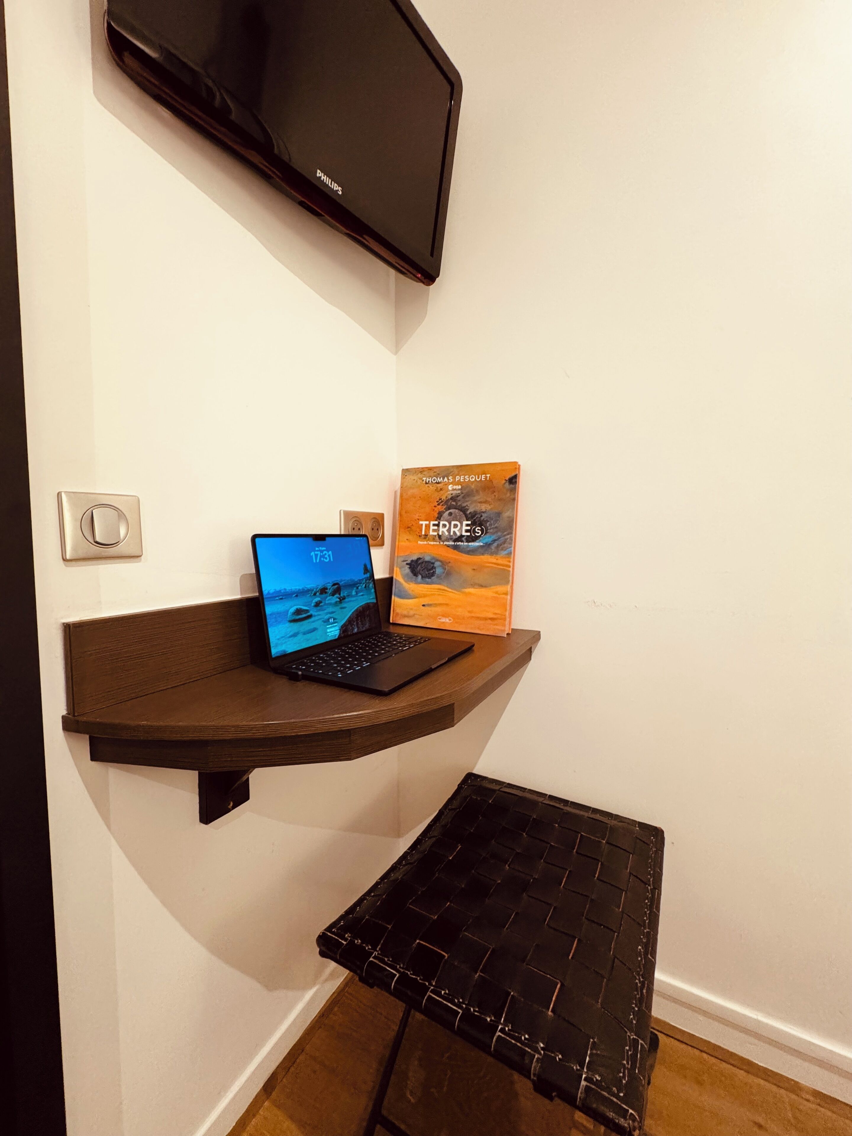 A corner desk with a laptop, a book titled 'Terre(s)', and a flat-screen TV mounted on the wall.