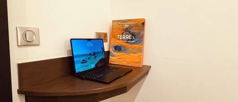 Un bureau d’angle avec un ordinateur portable, un livre intitulé « Terre(s) » et un téléviseur à écran plat fixé au mur.
