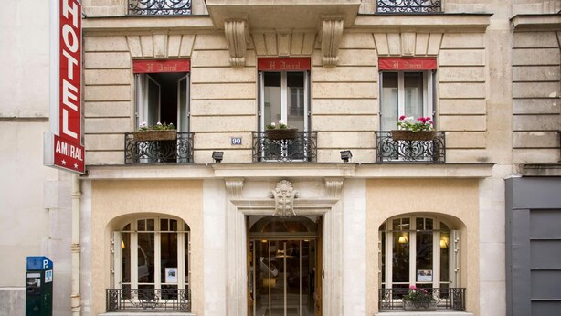 A hotel facade with a classic architectural style, featuring a prominent entrance with a glass door, arched windows, and decorative iron railings on the balconies. - Amiral (Paris)