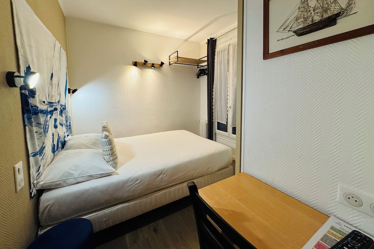 Une petite pièce, de taille modeste, comprenant un lit simple, une table en bois et une chaise. Un étagère est fixée au mur au-dessus du lit, et un tableau encadré est accroché au mur.