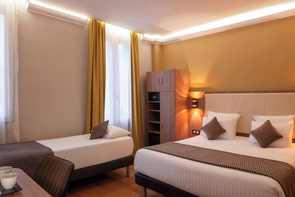 Une chambre d’hôtel avec deux lits simples, une armoire en bois, une petite table avec deux tasses et une fenêtre avec des rideaux jaunes.