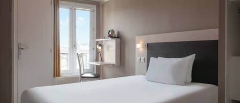 Une chambre d’hôtel avec un grand lit, une fenêtre avec des rideaux, une table de chevet et une étagère fixée au mur, sur laquelle sont posés divers objets.