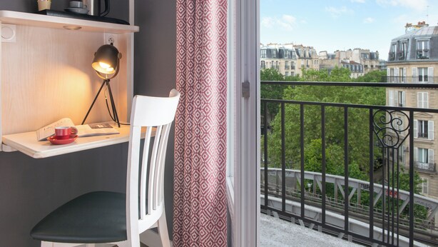 Un balcón con vista a edificios parisinos, una silla blanca, un escritorio con una lámpara y una taza roja.