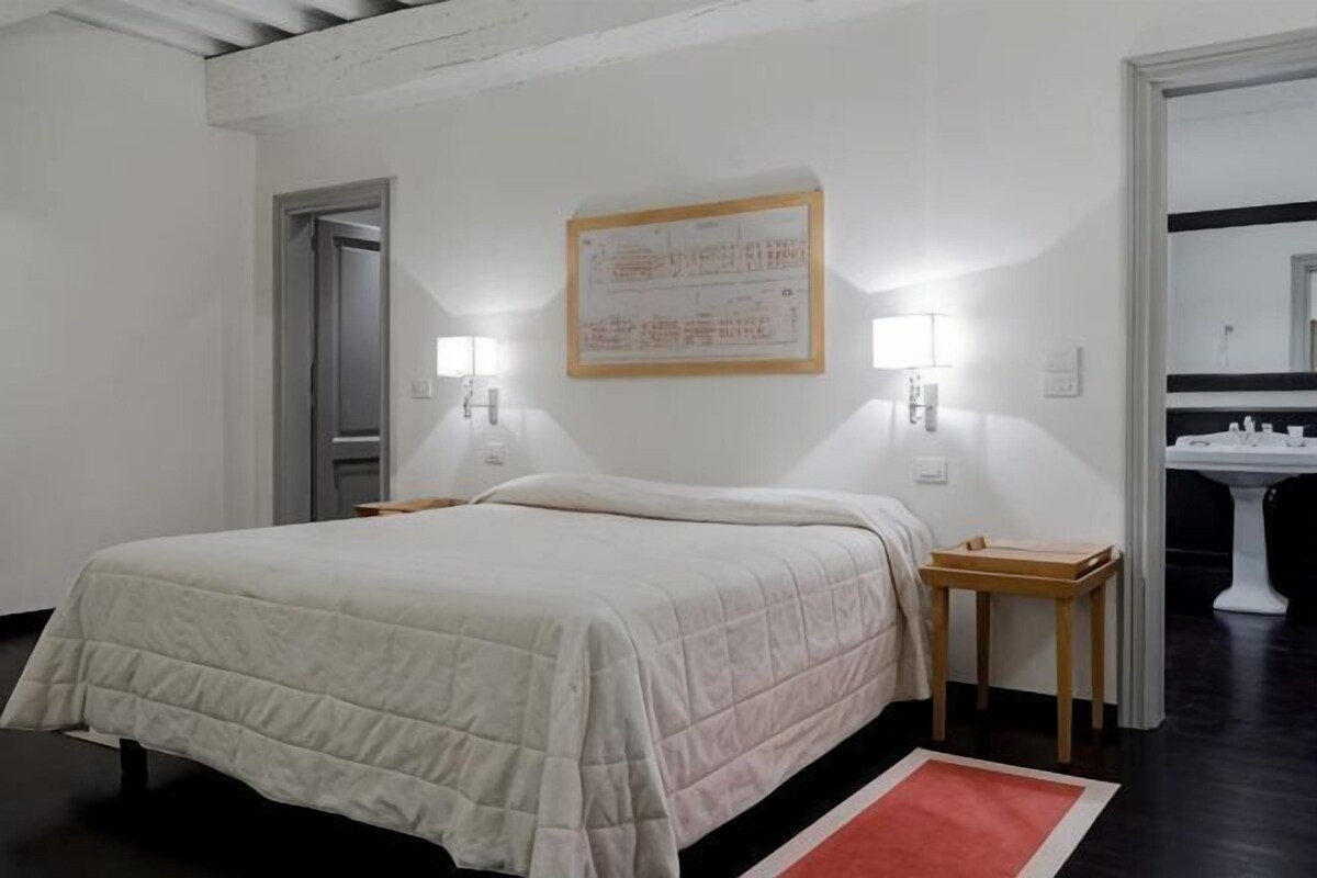 Une chambre d’hôtel comprenant un lit, des tables de chevet, une horloge murale et une salle de bain avec un lavabo et un miroir.