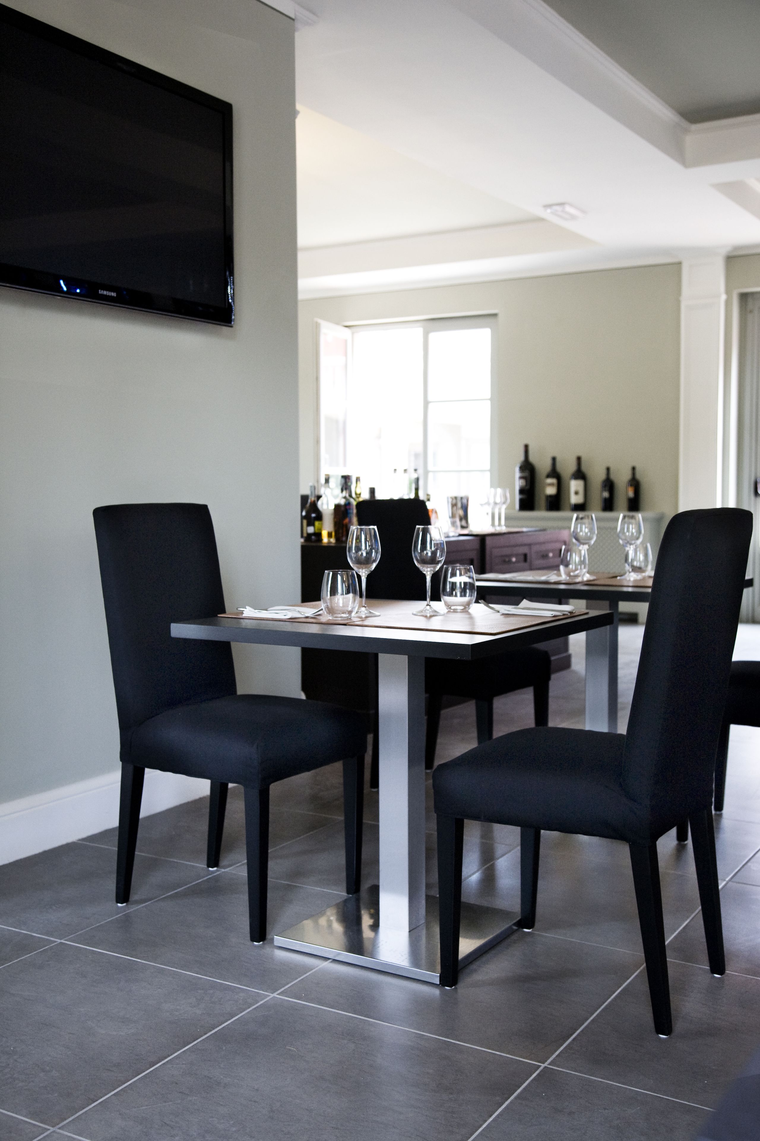 A modern dining area with a large flat-screen TV mounted on the wall, a dining table with four chairs, and a kitchen in the background with wine bottles on the counter.