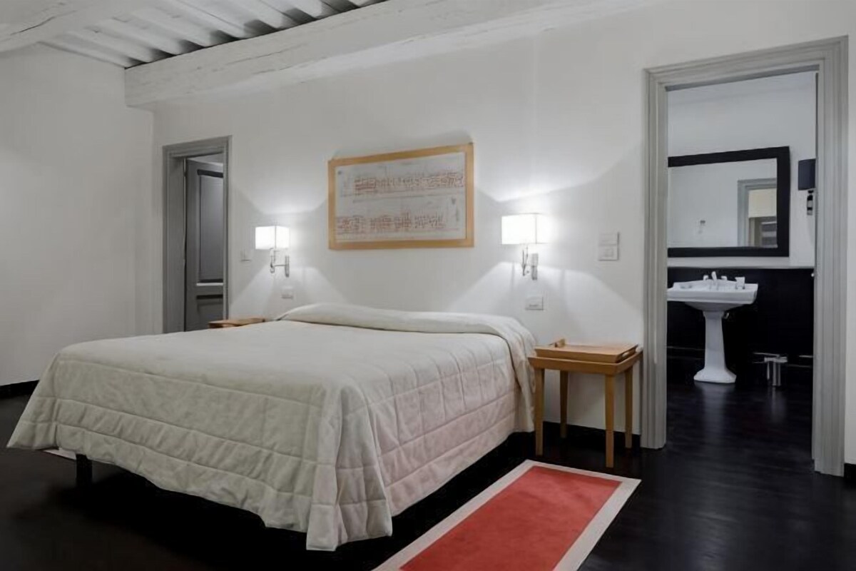 Une chambre à coucher avec un lit, des lampes de chevet, une table de chevet, un tapis rouge, un mur blanc orné d’un tableau encadré, un lavabo blanc et un miroir.