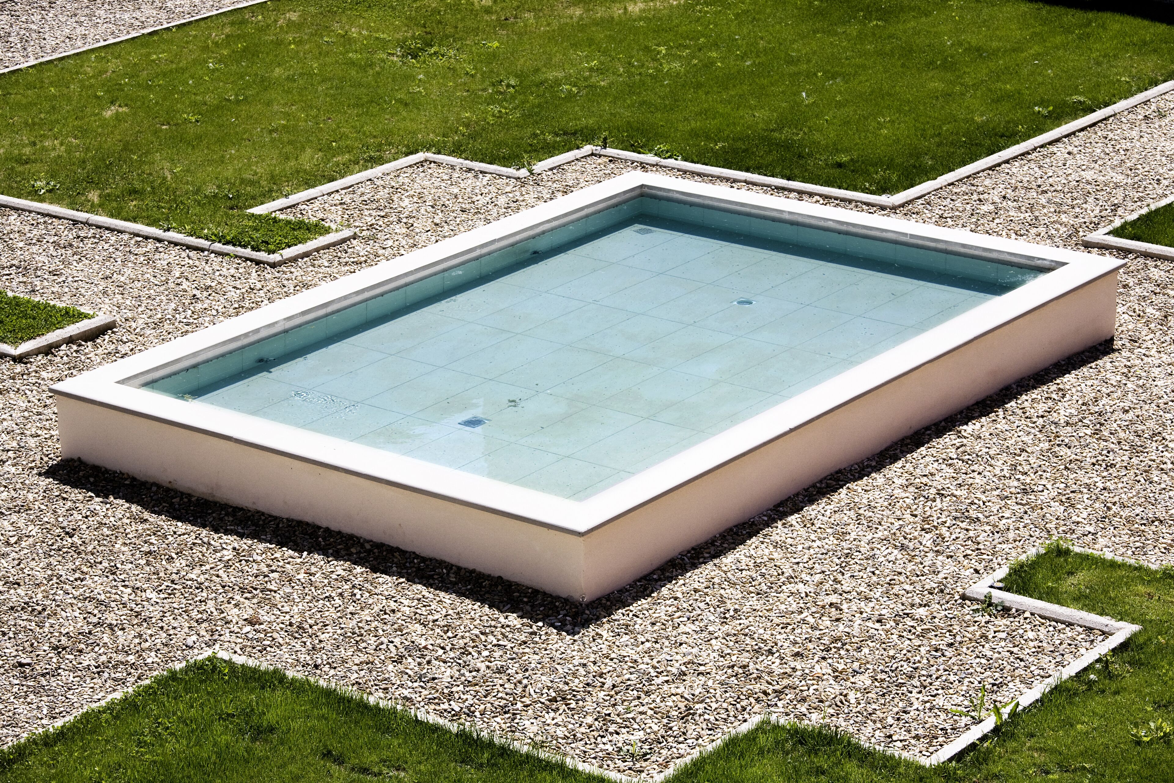 A rectangular pool with a white border and a tiled interior, surrounded by a gravel area and grass.