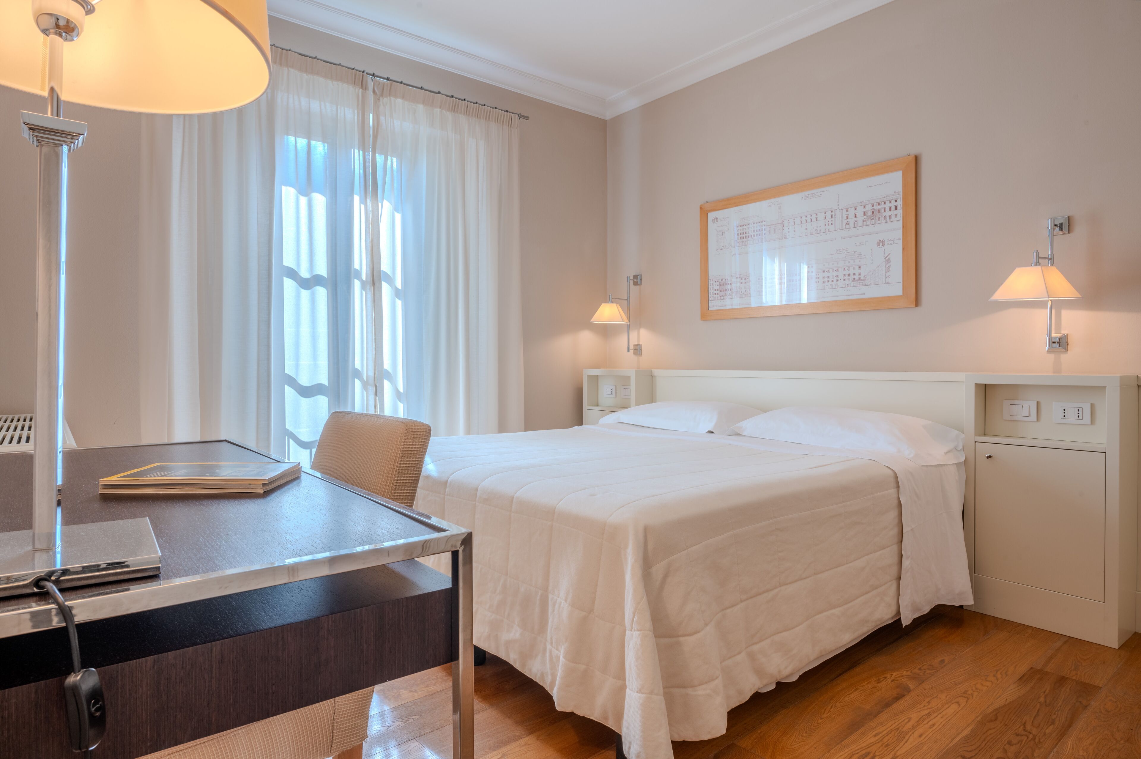 Een hotelkamer met een bed, bureau, stoel, lamp en een lijst met een architectuurtekening aan de muur.