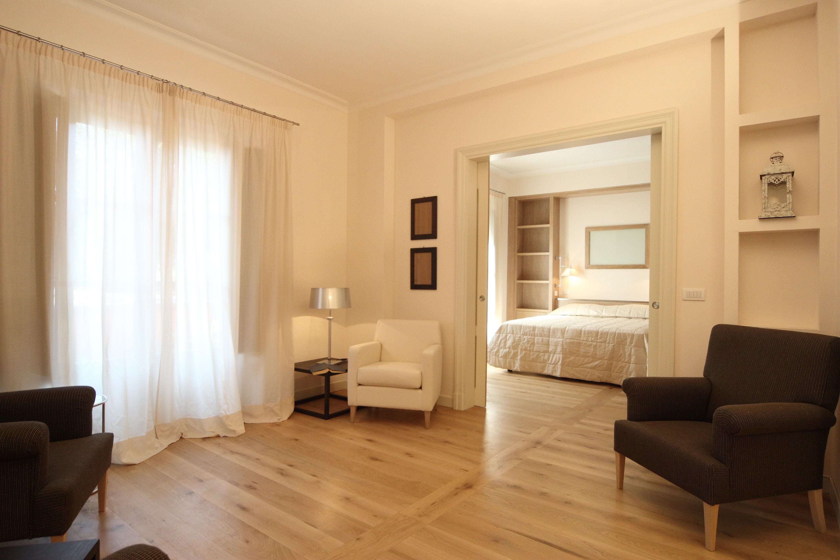 A hotel room with a wooden floor, a bed with a white duvet, a white armchair, a dark armchair, and a small table with a lamp.