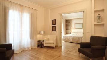 A hotel room with a wooden floor, a bed with a white duvet, a white armchair, a dark armchair, and a small table with a lamp.