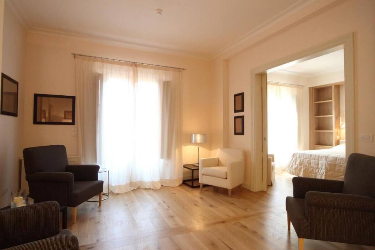 Habitación de hotel con piso de madera, dos sillones, una mesita de noche con una lámpara, una cama con cubrecama blanco y una puerta que da a un baño.