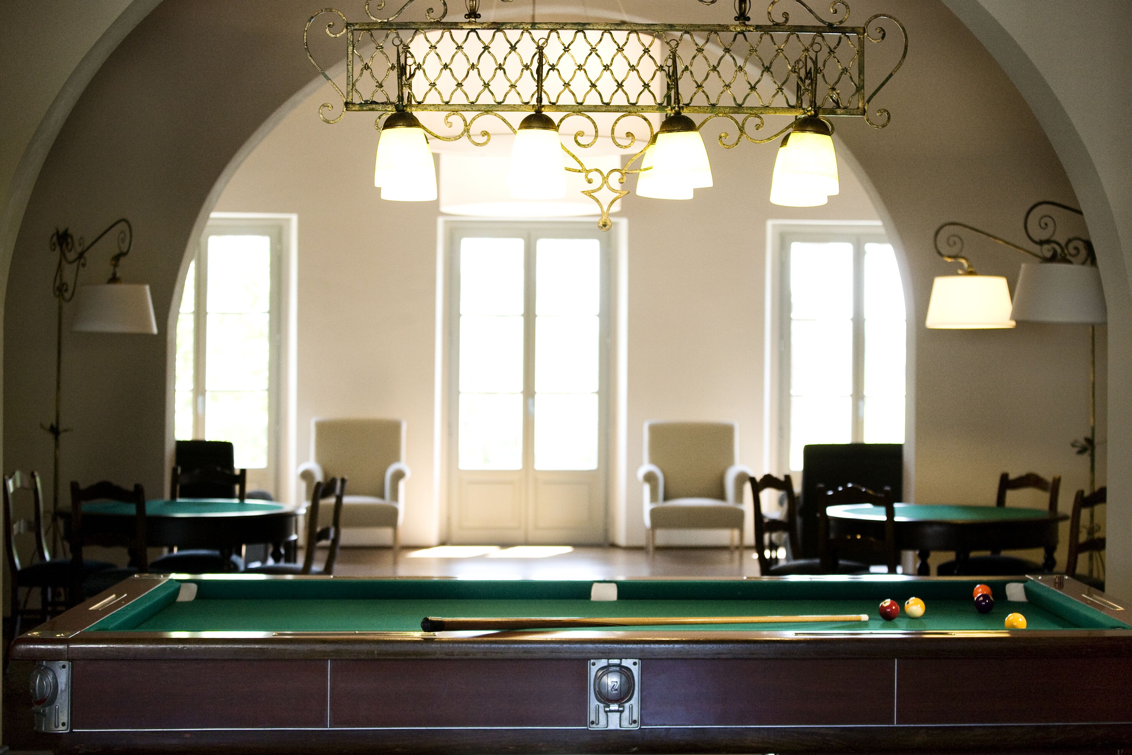 A pool table with balls, chairs, and a lamp in a room with arched ceilings and windows.