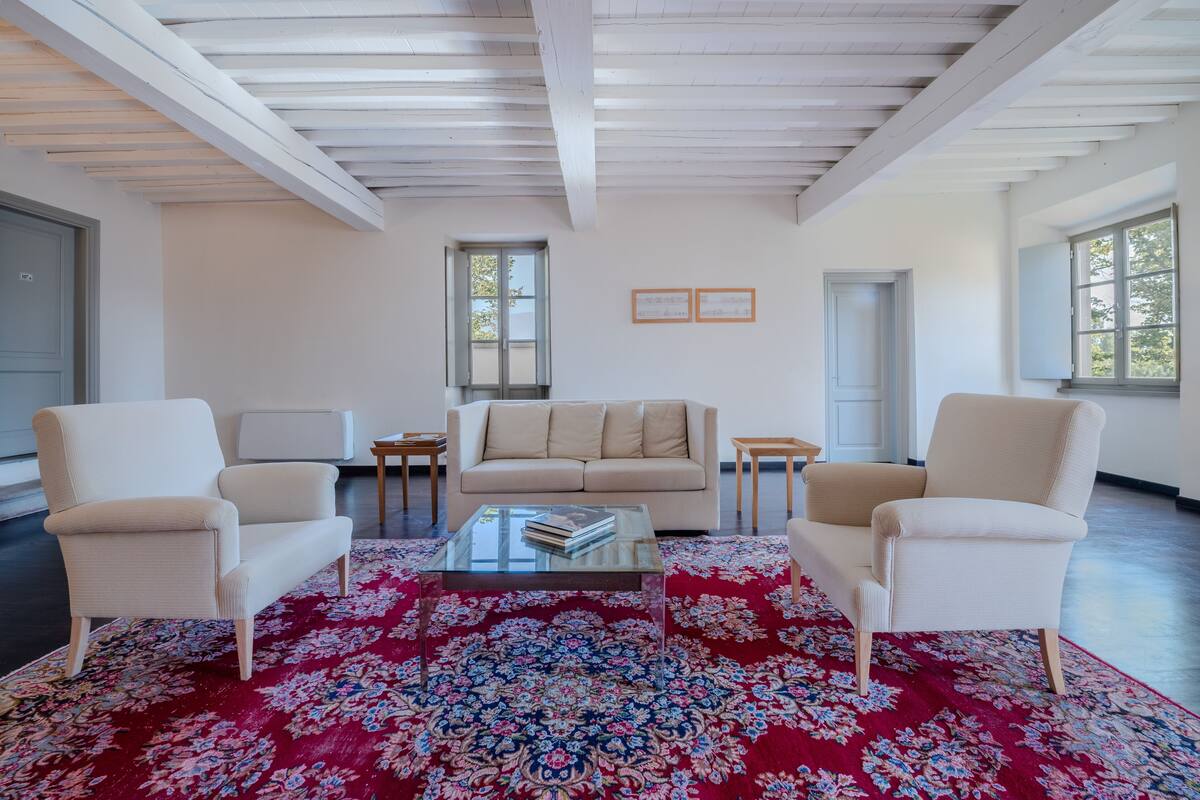 Amplia sala de estar con un tapete floral rojo grande, un sofá beige y dos sillones. Hay dos mesitas auxiliares y una mesa de centro de vidrio. La habitación tiene paredes blancas, techo de madera y ventanas grandes que dejan entrar luz natural.