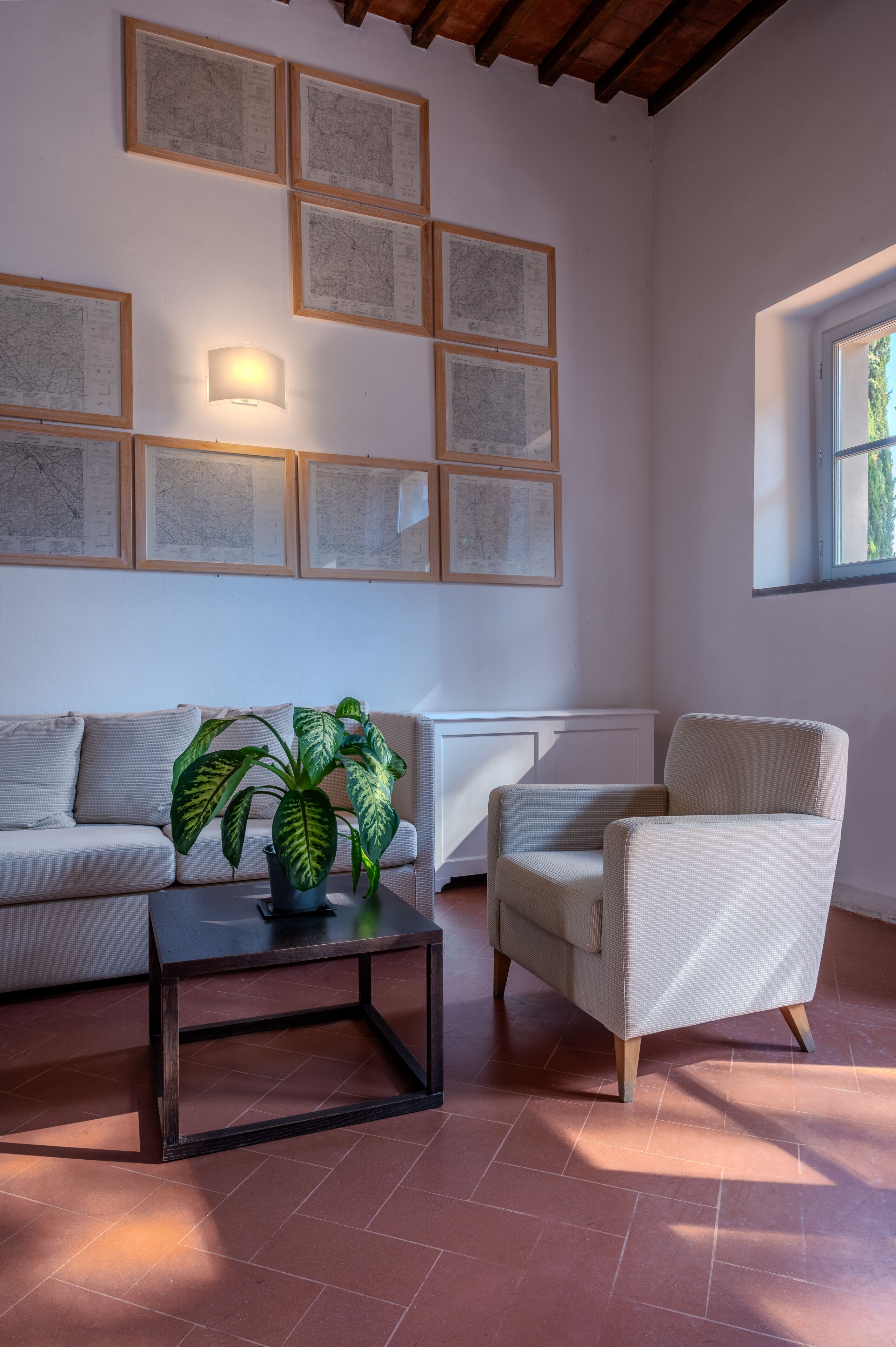 Una sala de estar con sofá, sillón, mesa de centro y planta en maceta. Hay mapas enmarcados en la pared y una ventana con luz natural.