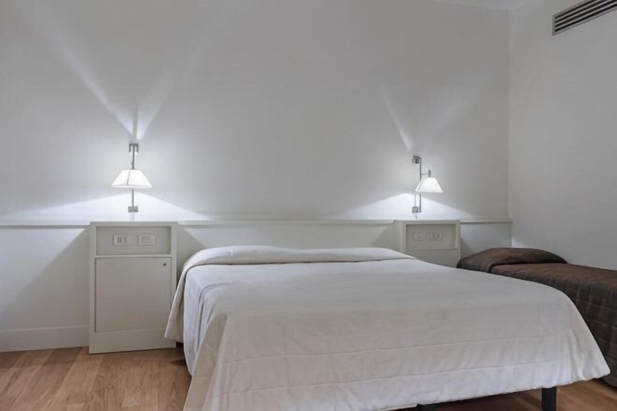 Une chambre à coucher comprenant un lit, deux tables de chevet, une petite table et une bouche d’aération fixée au mur.