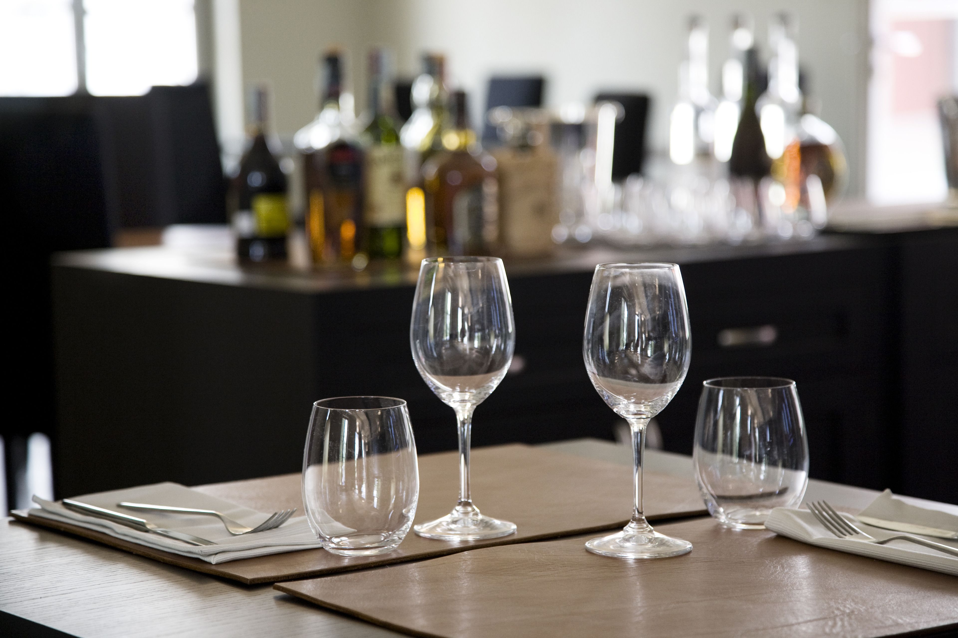 A table set with three wine glasses and cutlery, with bottles in the background.