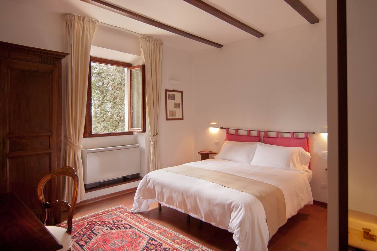 Un dormitorio con una cama grande, cabecera de madera, una ventana con cortinas, una silla de madera y un tapete rojo estampado.