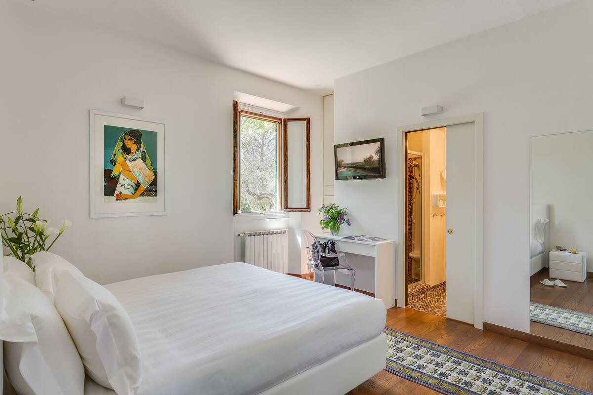 Un dormitorio con cama, escritorio y silla. Se ve una ventana, un radiador y una puerta que da a otra habitación.