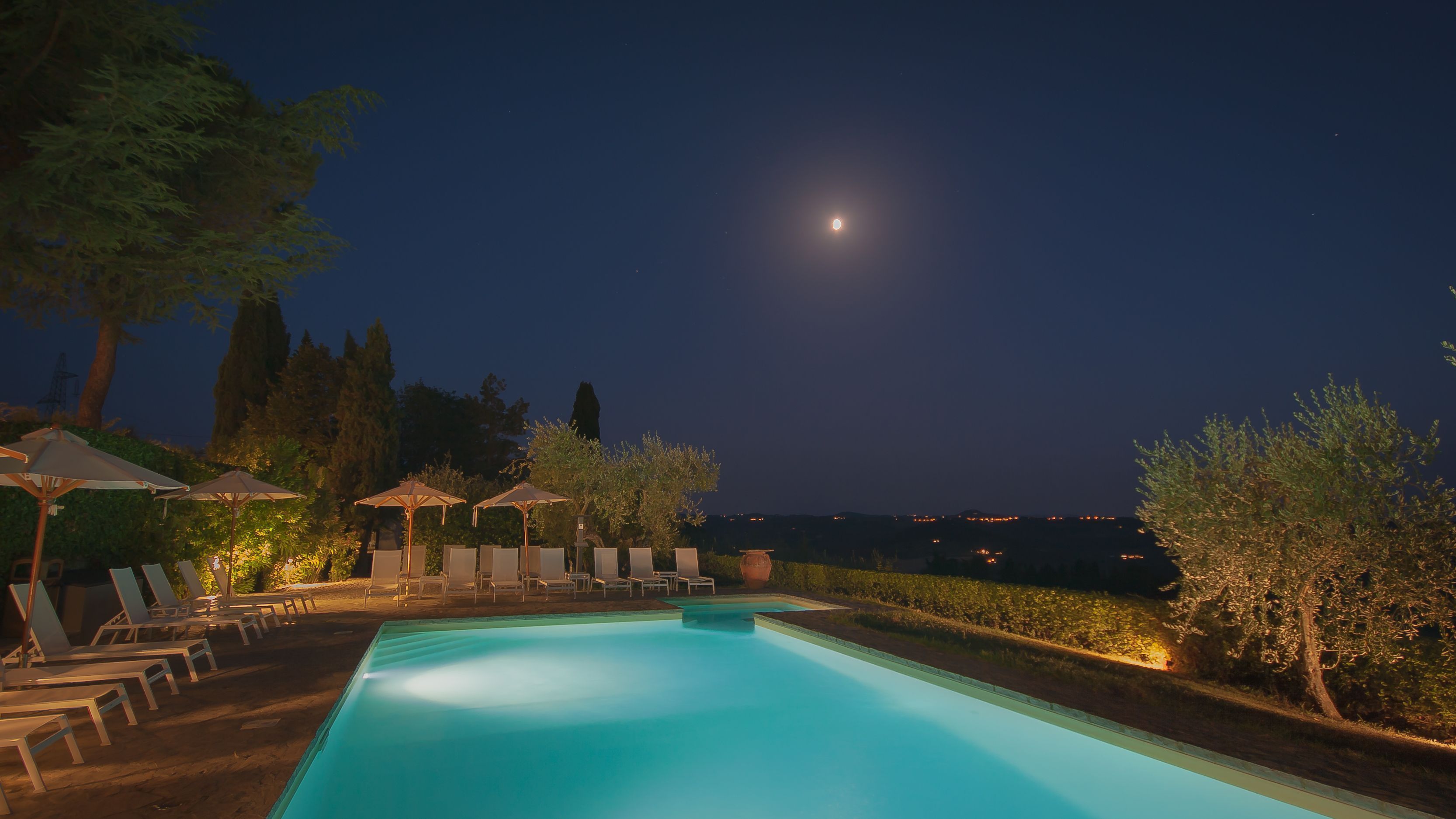 A nighttime pool area with lounge chairs and umbrellas, surrounded by trees and a view of the city skyline.