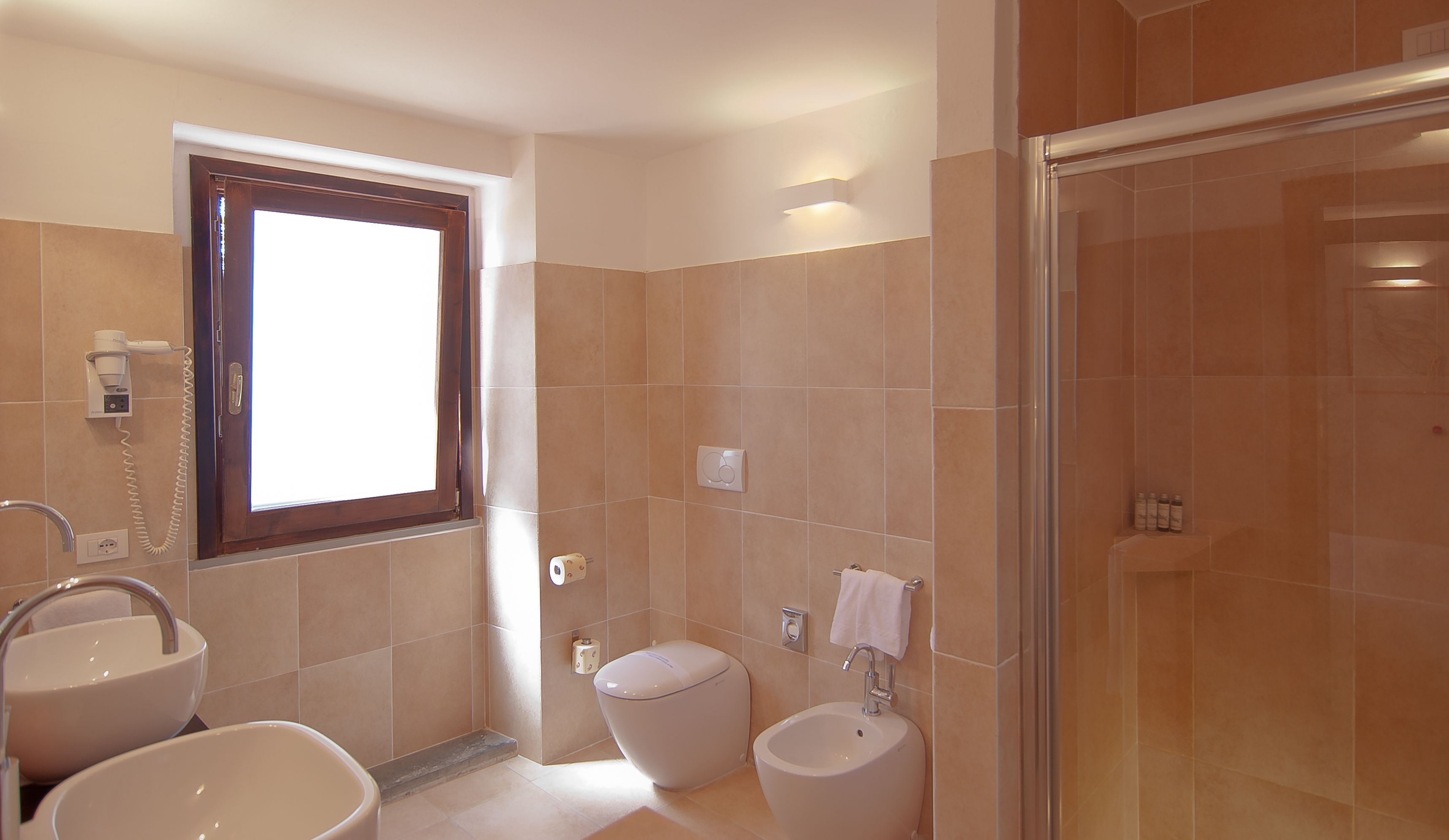 Une salle de bain comprenant des toilettes, un bidet et un lavabo. Il y a une fenêtre avec un cadre en bois et un téléphone fixé au mur.
