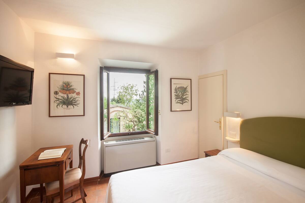Una habitación de hotel con una cama, un escritorio con una silla, una ventana e impresiónes botánicas enmarcadas en las paredes.