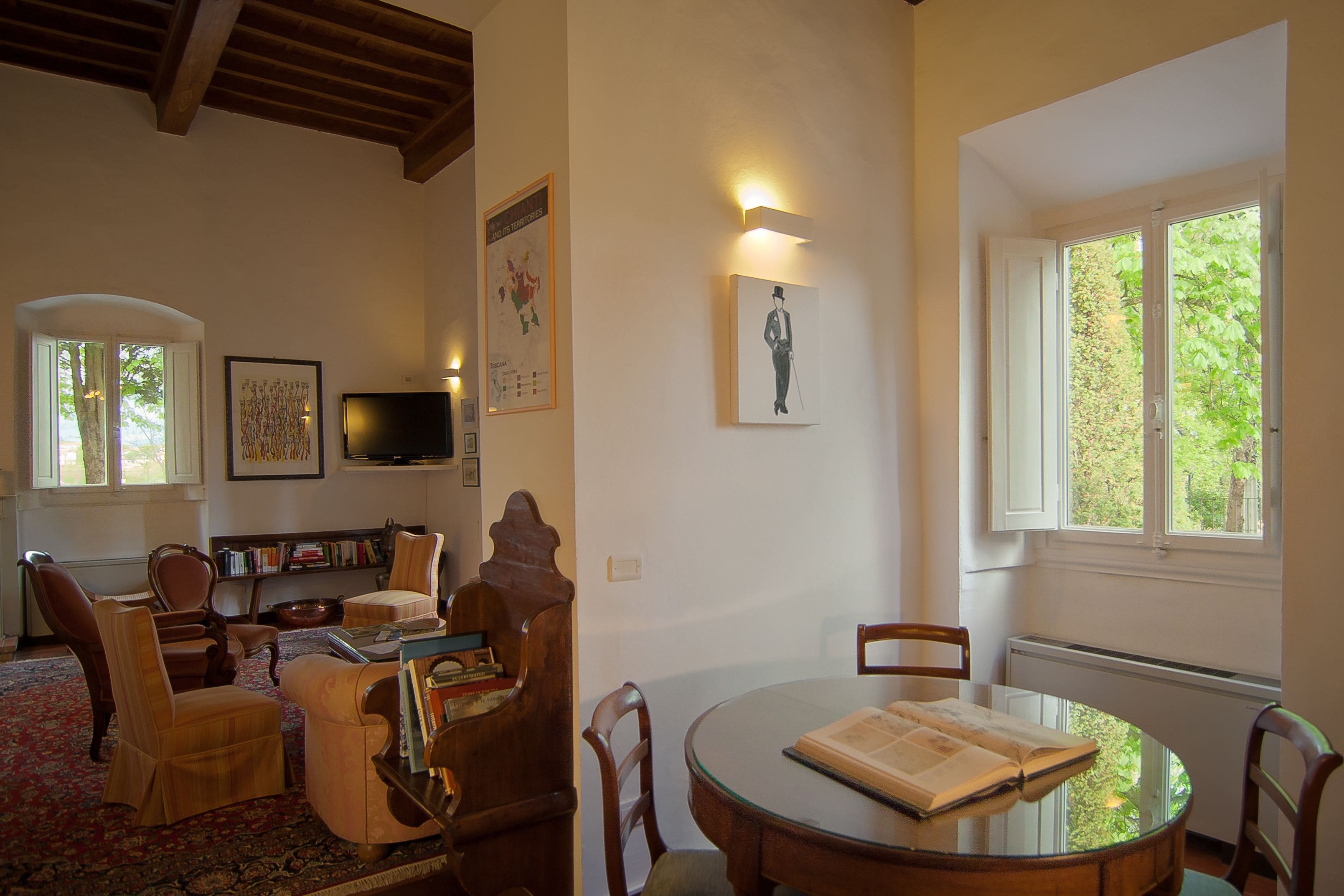 Una sala de estar acogedora con una mesa redonda de vidrio, sillas de madera, una estantería y un televisor.