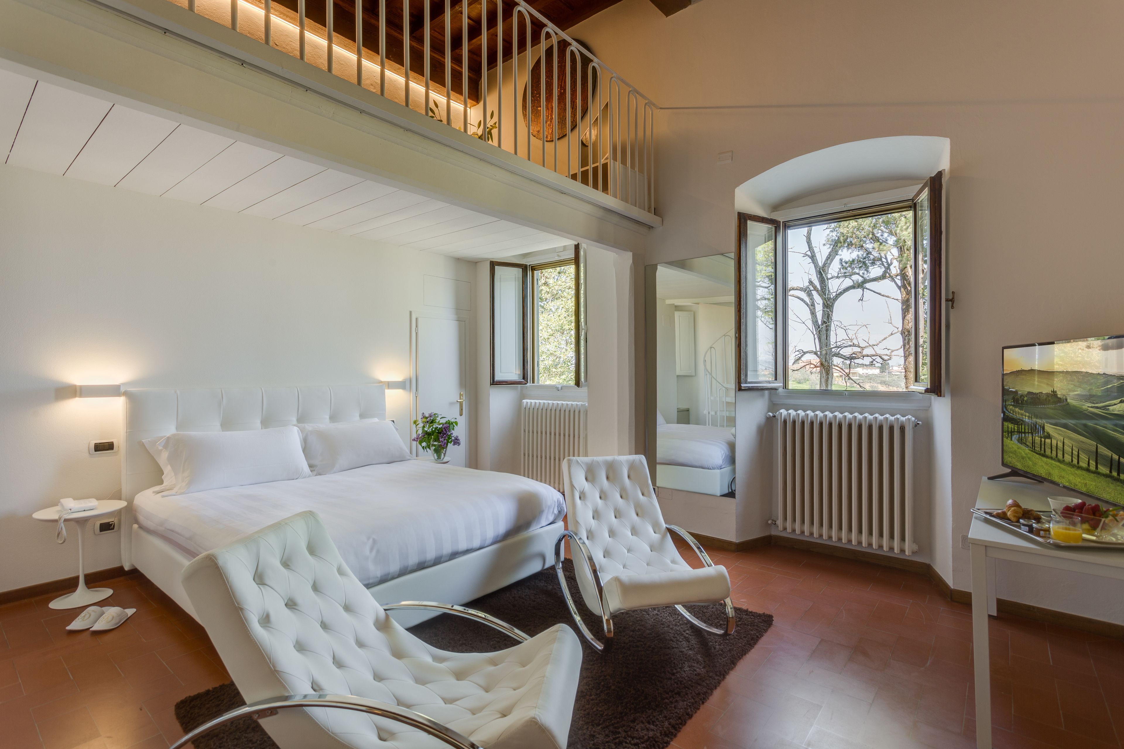 Un dormitorio moderno con una cama grande, dos sillones blancos, una mesita con provisiones para el desayuno, un televisor y vistas al exterior.