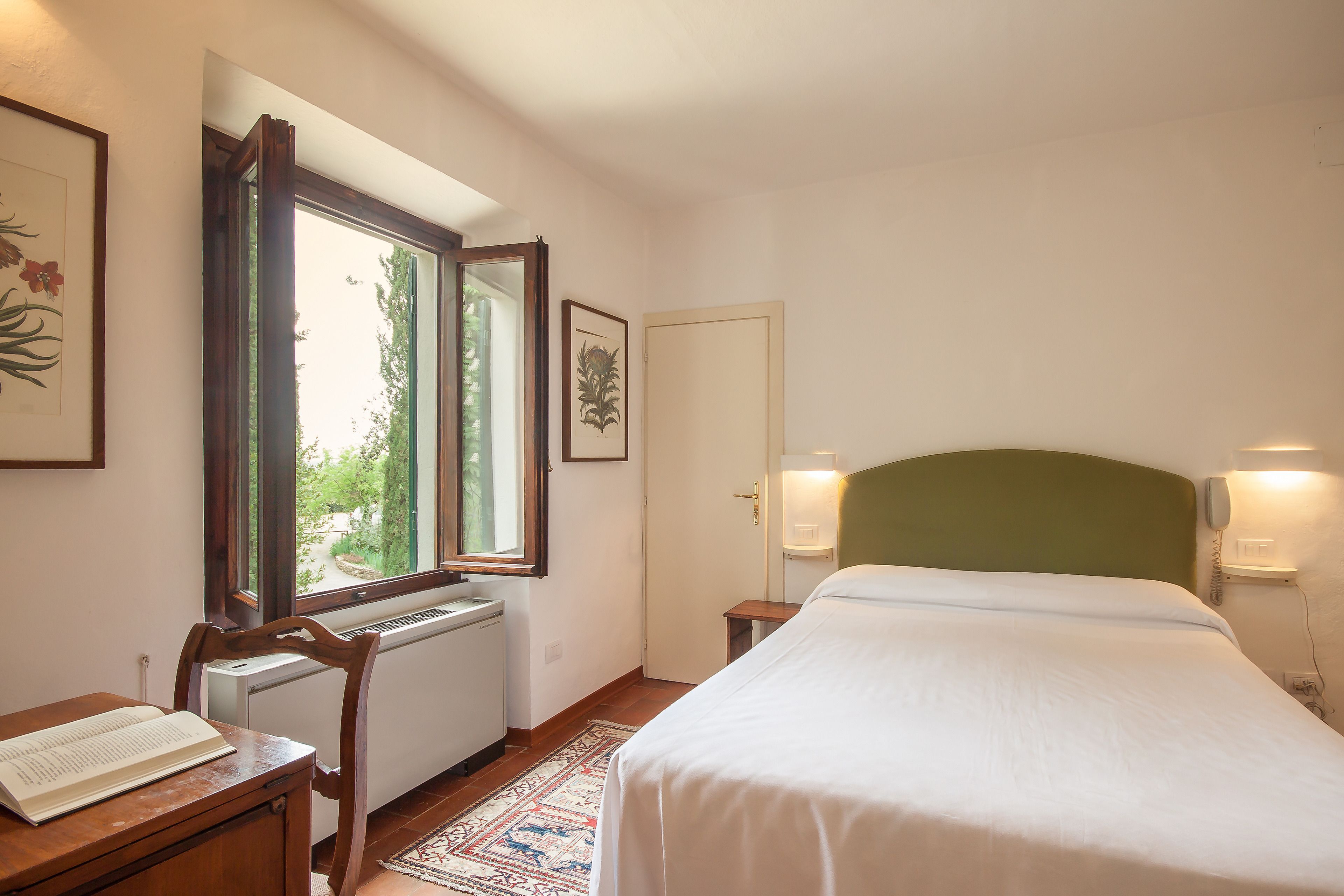 A bedroom with a bed, a desk with a book, a radiator, a window, and a door.