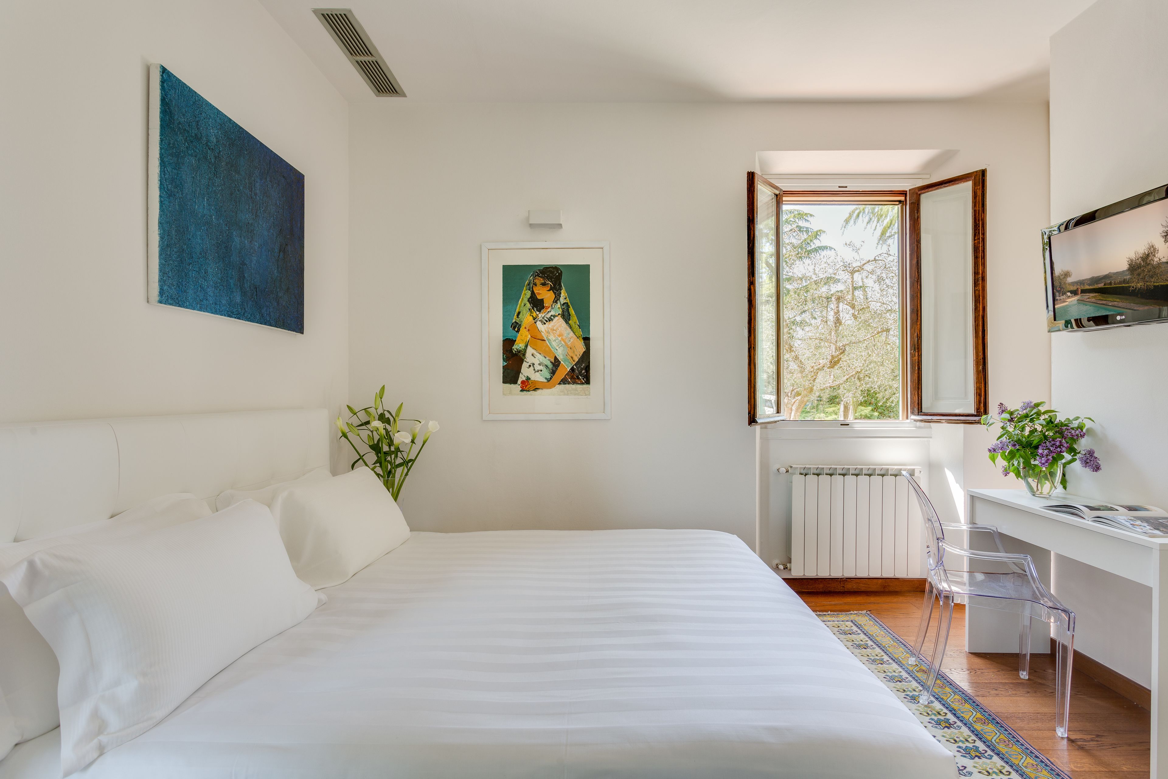 A bedroom with a large bed, a desk with a chair, a window, and artwork on the walls.