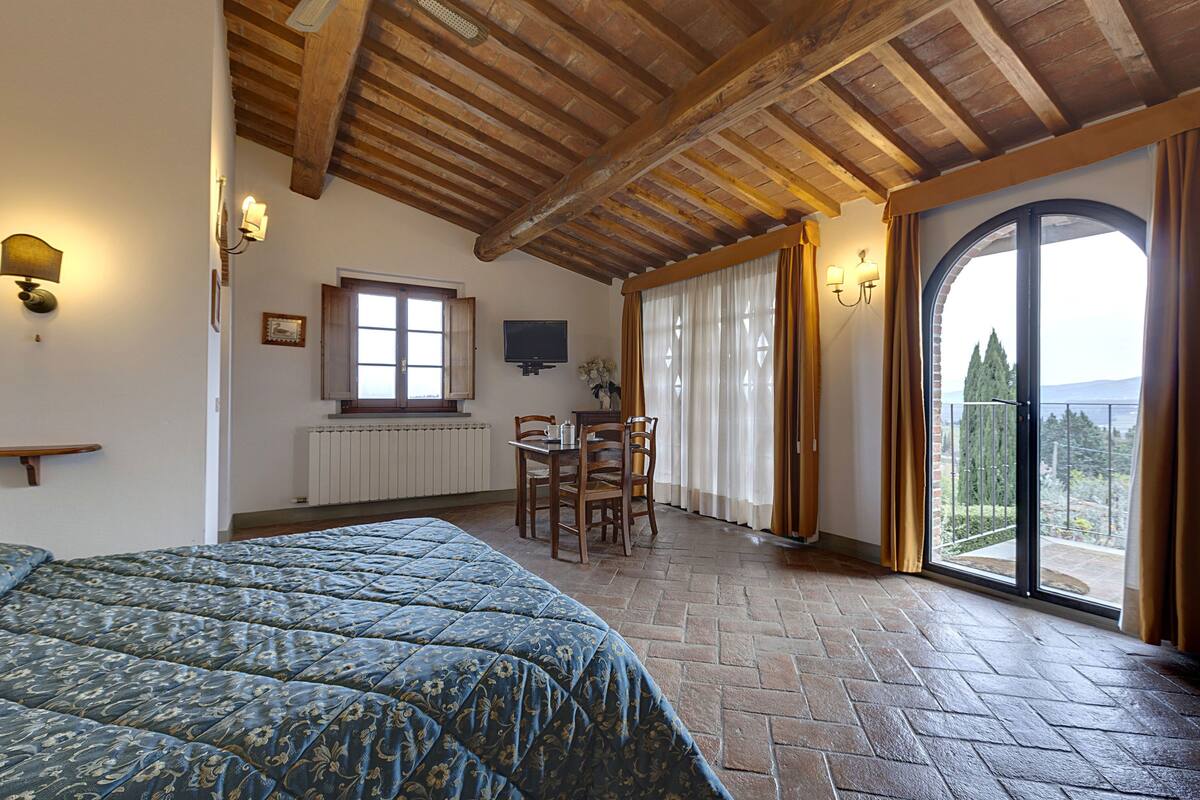 Un dormitorio con una cama grande, techo de madera, una mesa de comedor con sillas, televisor y un balcón con cortinas.