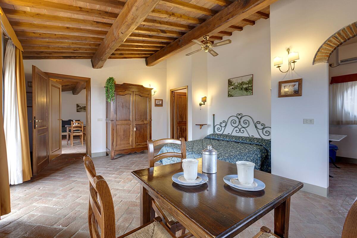 Un comedor con una mesa de madera dispuesta para dos personas, un ventilador de techo y una cama con colcha estampada al fondo.