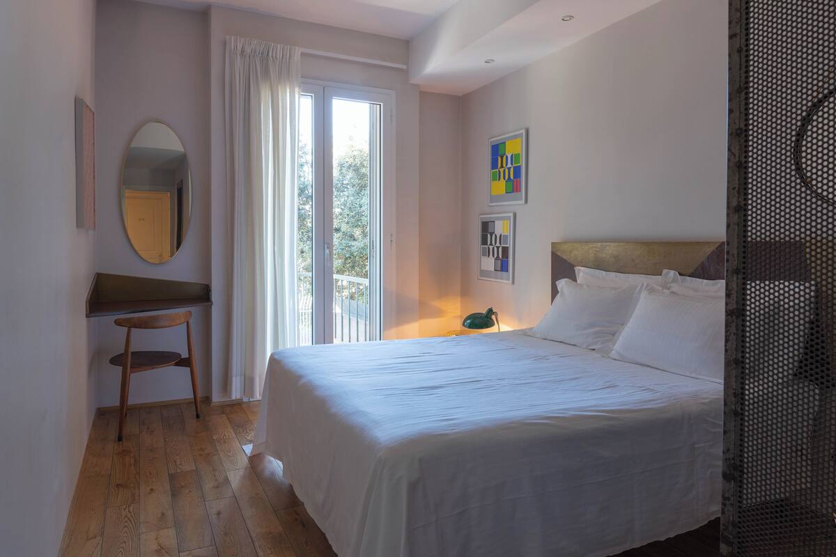 A bedroom with a bed, a desk, a chair, a window with curtains, and a wall with framed pictures.