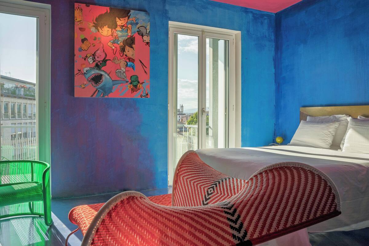 A room with a bed, a green chair, a red and white patterned chair, a large window, and a colorful wall art piece.
