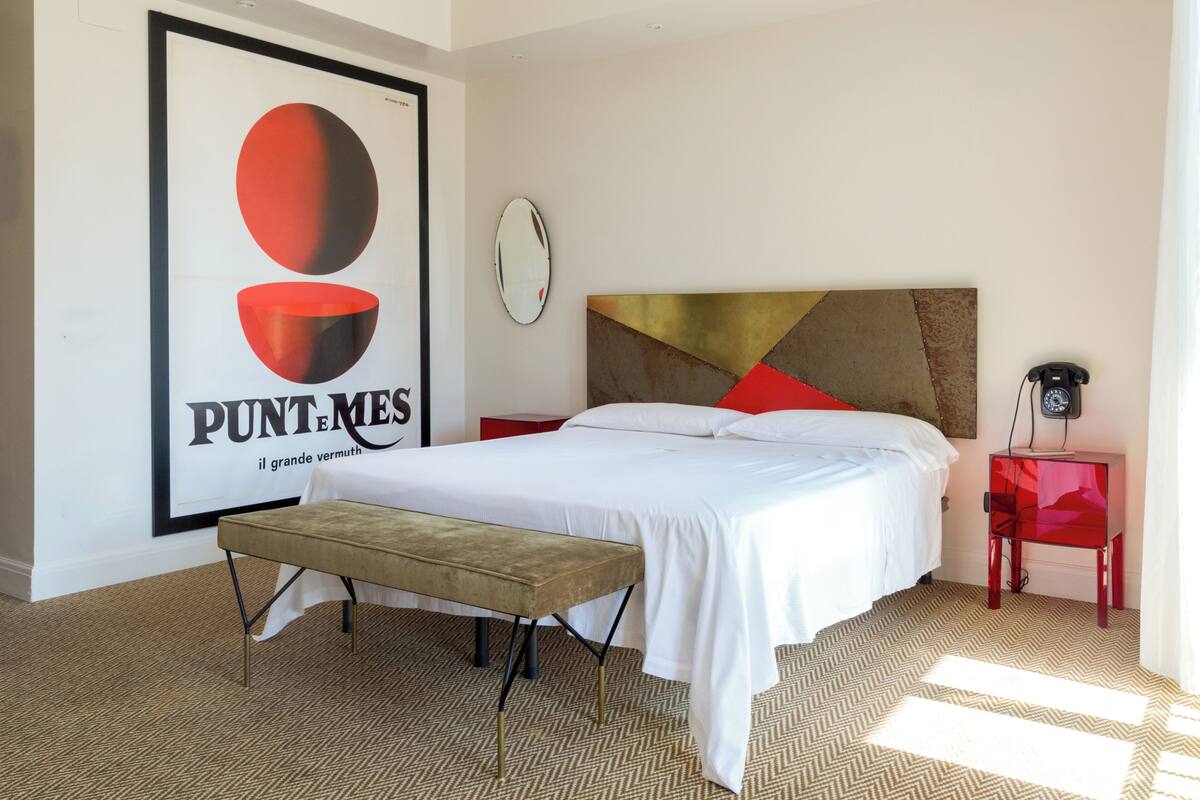 A modern bedroom with a large bed, a bench, a red accent wall, and a poster on the wall.