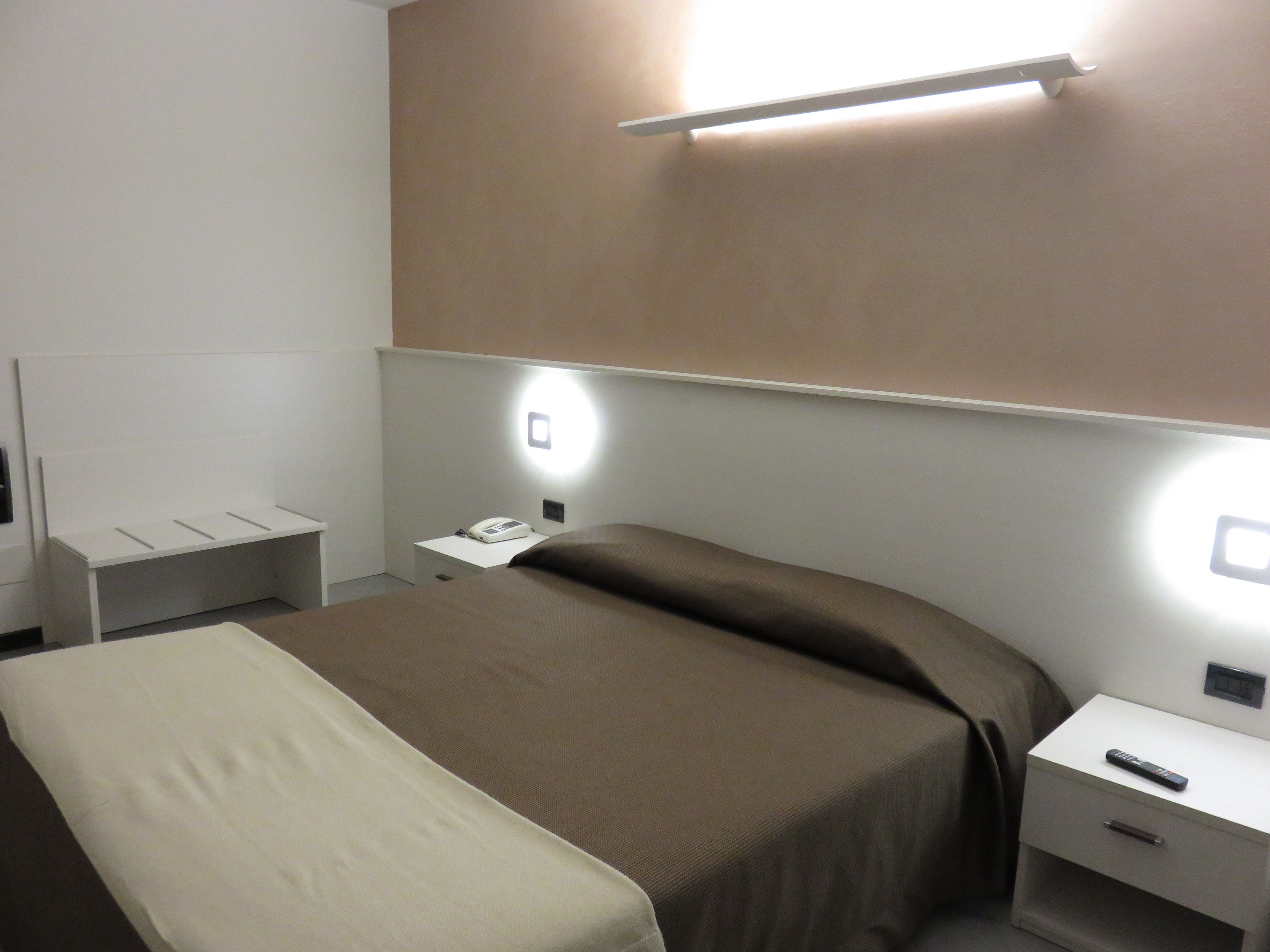 Habitación de hotel con cama, mesita de noche con teléfono, control remoto e iluminación en la pared.