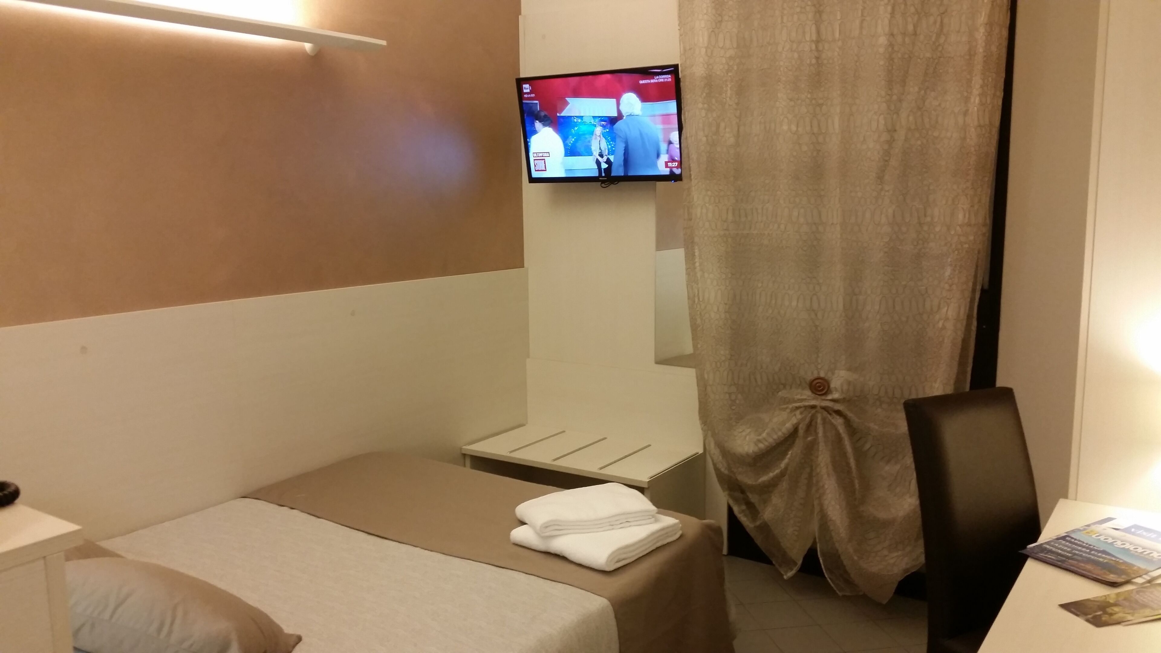 Une chambre d’hôtel avec un lit simple, une petite table avec une chaise, un téléviseur fixé au mur et un rideau.