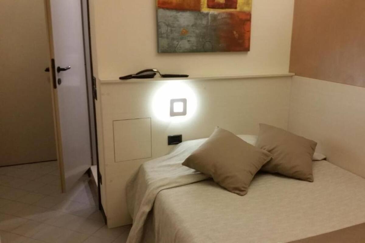Un dormitorio pequeño y ordenado con una cama, dos almohadas, una mesita de noche con una lámpara y un cuadro en la pared.