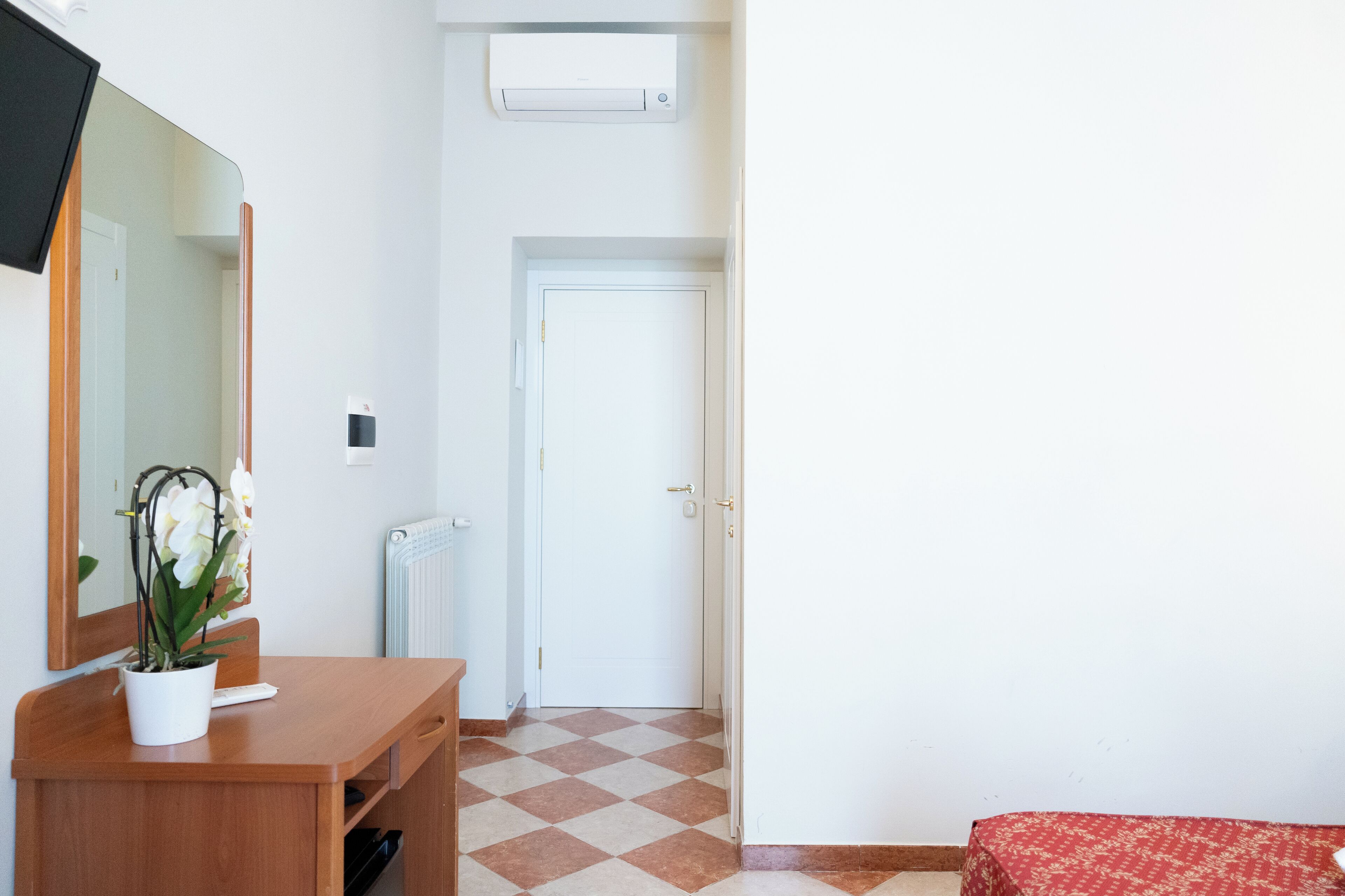 A hotel room with a wooden desk, a potted orchid, a mirror, a radiator, a checkered floor, a white door, and a red patterned bedspread.