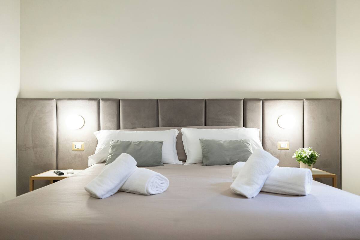 A neatly made bed with pillows, a remote control, and a small plant on a bedside table.