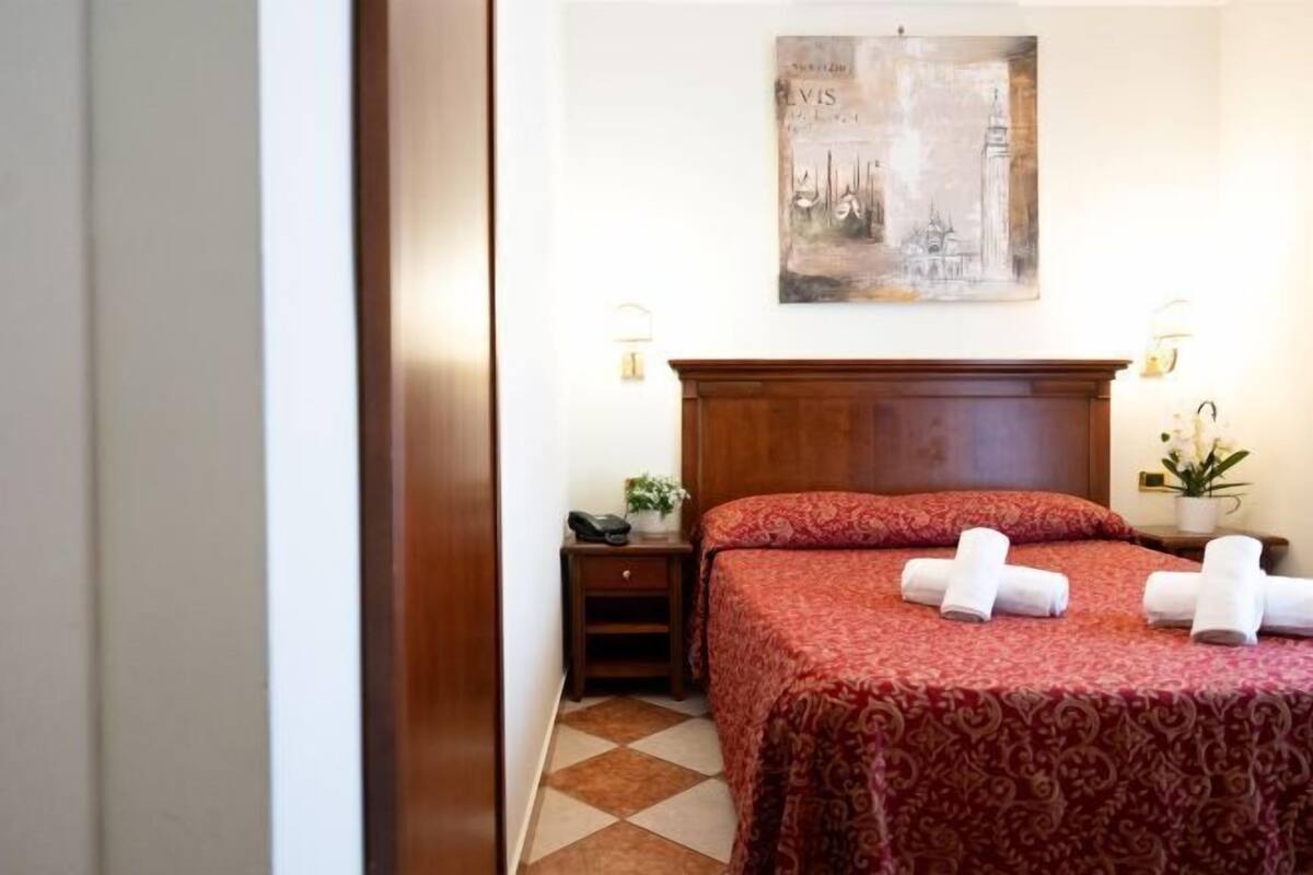 A hotel room with a bed, a nightstand, a lamp, a painting on the wall, and a tiled floor.